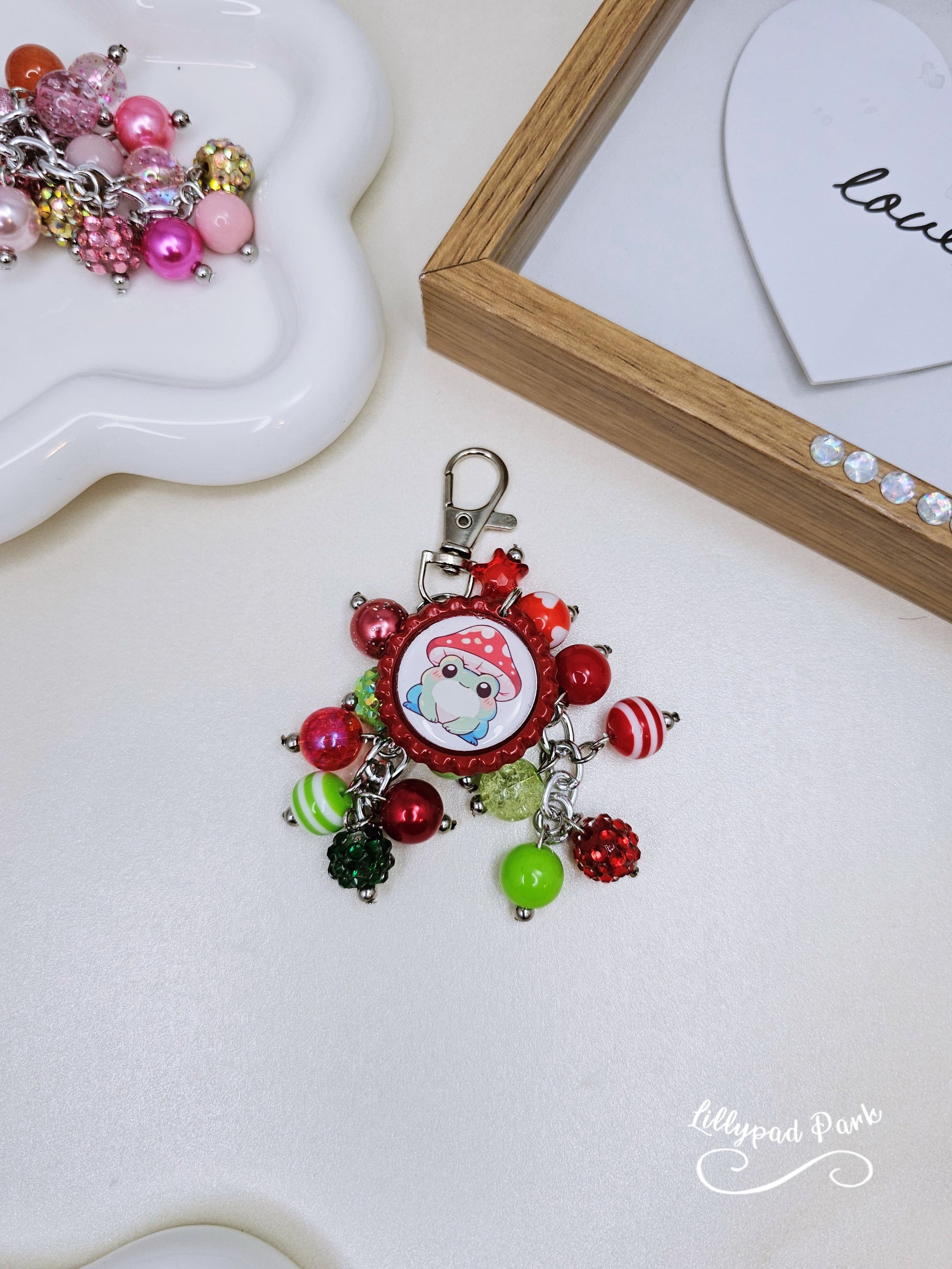 A colorful beaded bag charm featuring a whimsical frog wearing a mushroom cap, with red and green beads and a bottle cap charm.