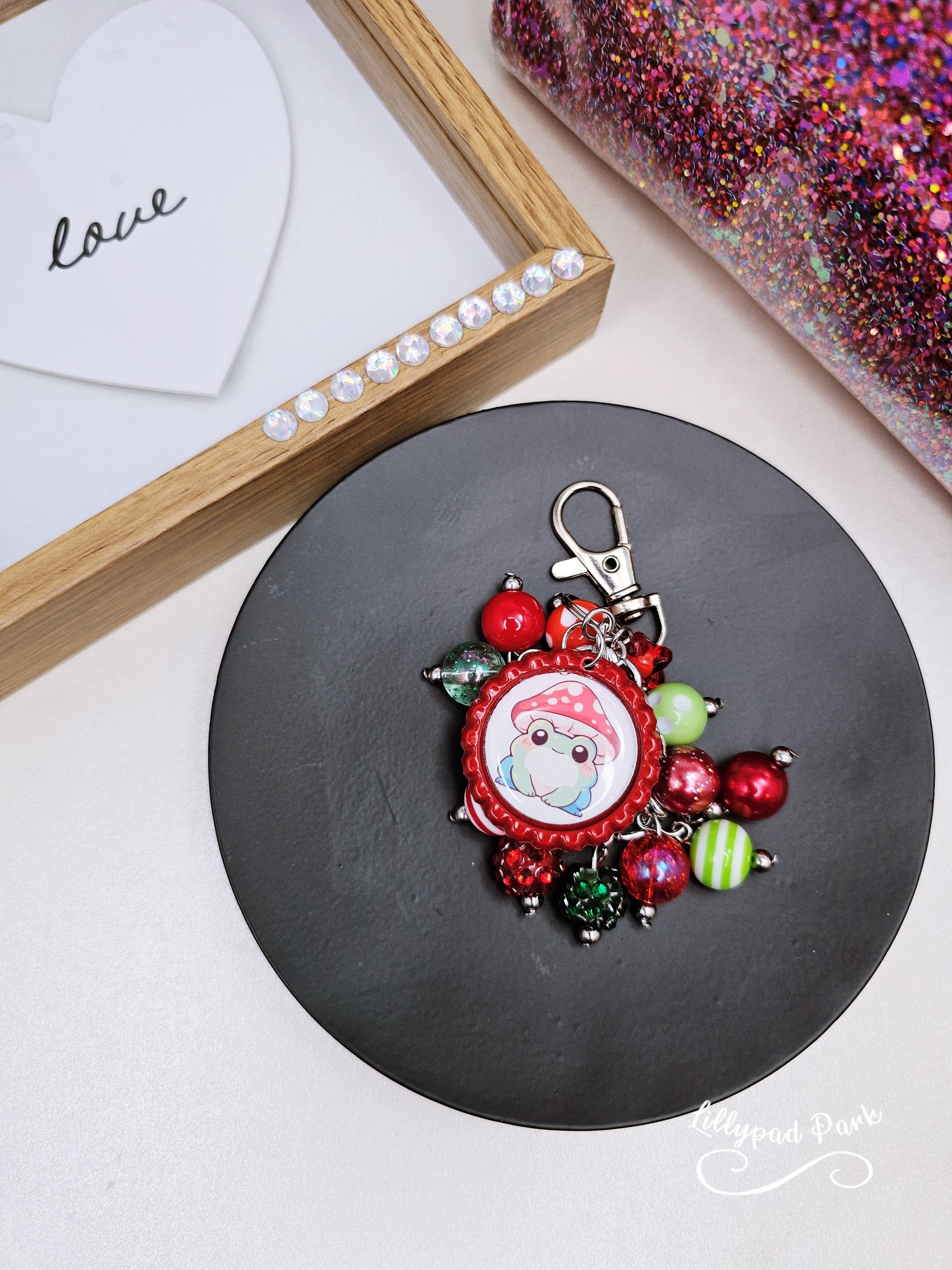 A colorful beaded bag charm featuring a whimsical frog wearing a mushroom cap, with red and green beads and a bottle cap charm.