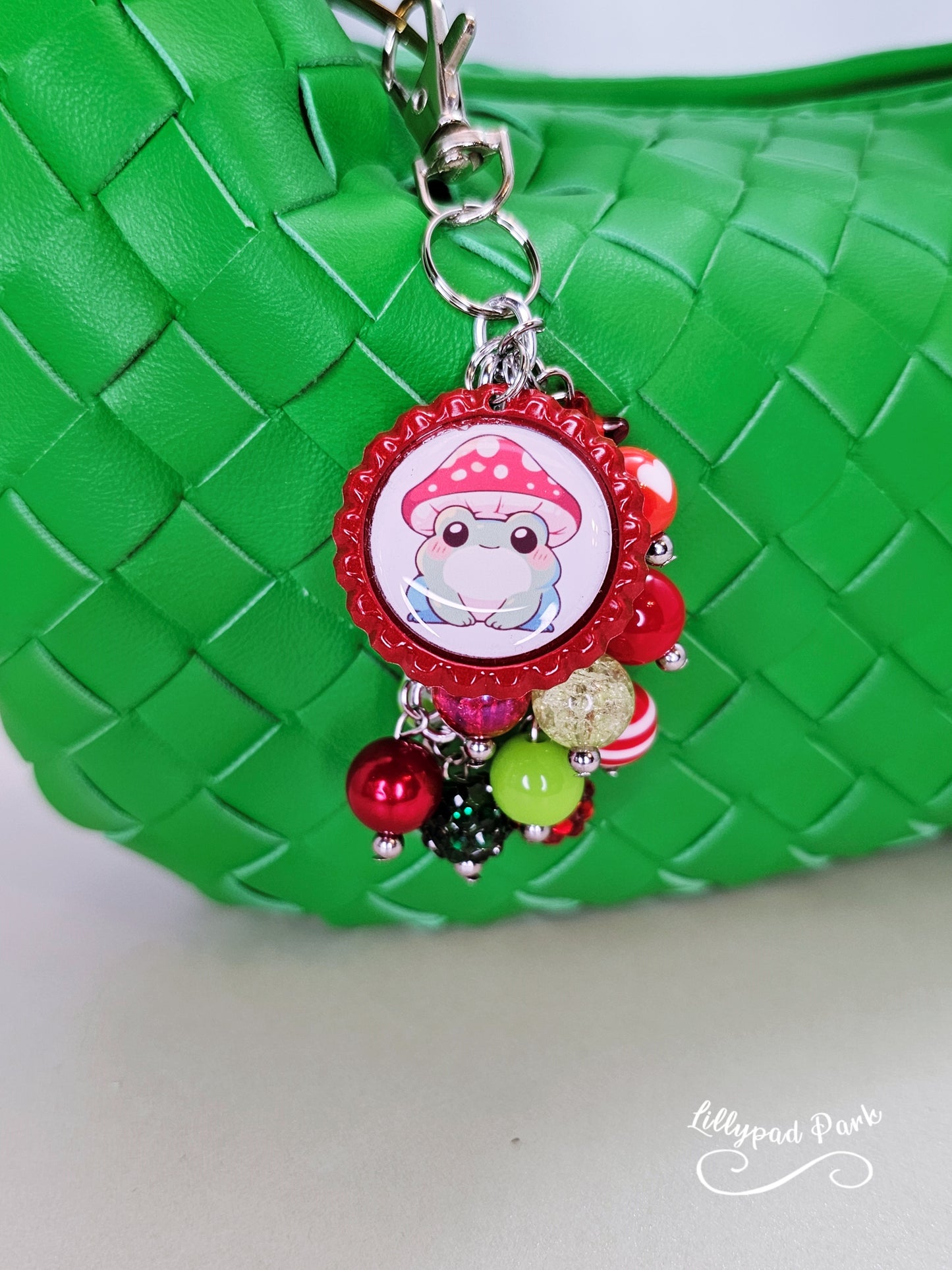 A colorful beaded bag charm featuring a whimsical frog wearing a mushroom cap, with red and green beads and a bottle cap charm.